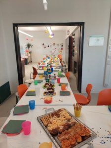 Viele aneinander gestellte Tische mit Stühlen dran. Auf den Tischen stehen bunte Becher und Teller mit Servierten. Essen ist auf den Tischen verteilt. Die Tafel mit Tischen geht durch eine große offene Tür durch. 