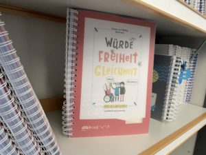 Ausschnitt eines Bücherregals aus der Schulbücherei. Die meisten Bücher sind ordentlich ins Regal gestellt. Ein Buch steht frontal mit dem Cover nach vorne. Es heißt "Würde, Freiheit, Gleichheit".