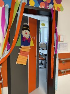 Tür zu einem Raum, die halb offen steht. Oberhalb von der Tür sind mit Krepppapier Fänden befestigt, die wie ein zur Seite gebundener Vorhang hängen. Auf dem Türblatt ist ein Clown aus Pappe geklebt.