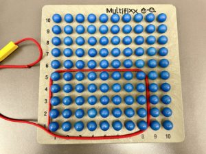 Holzbrett mit 100 in 10x10 Feld angeordneten aufgeklebten Kugeln in Blau. Um Die Kugeln aus den Reihen 1-5 waagrecht und 1-8 senkrecht ist eine rote Schnur gelegt. 
