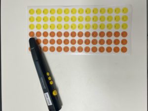 Schwarzer dicker Stift mit vier gelben Punkten auf der Seite. Der Stift liegt auf in Reihe geklebten gelben und roten Punkten. 