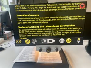 Bildschirm, auf dem Text in gelb auf schwarzem Hintergrund zu sehen ist, darunter liegt auf einem verschiebbaren Pult eine Zeitung. 