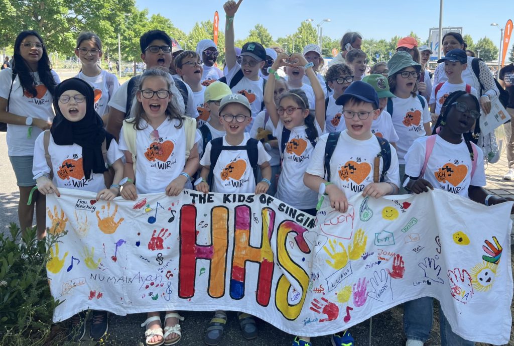 Auf dem Bild ist eine große Gruppe von Kindern und einigen Erwachsenen zu sehen, die in der Natur stehen und ein langes, weißes Banner hochhalten. Die meisten Personen auf dem Foto tragen weiße T-Shirts mit demselben Aufdruck und helle Kappen. Einige Kinder tragen Rucksäcke. Sie blicken lachend oder schreiend in die Kamera. Das von der Gruppe gehaltene Banner ist mit vielen bunten Handabdrücken, kleinen Zeichnungen und Namen versehen. In der Mitte sind die orangefarbenen Buchstaben „HHS“ zu sehen, die in einer geschwungenen Schriftart dargestellt sind. Darüber steht in Anführungszeichen: "THE KIDS OF SINGING". Im Hintergrund sind Bäume und eine asphaltierte Straße mit Pollern zu erkennen.