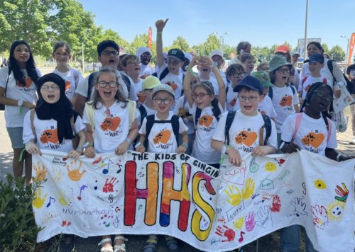 Auf dem Bild ist eine große Gruppe von Kindern und einigen Erwachsenen zu sehen, die in der Natur stehen und ein langes, weißes Banner hochhalten.